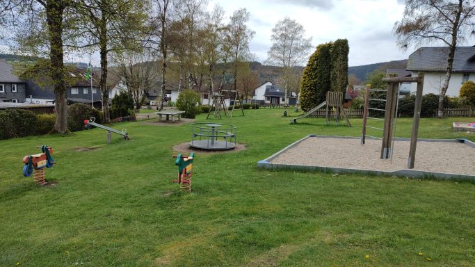 Spielplatz mit Rutschen, Schaukeln und grüner Wiese, umgeben von Bäumen.